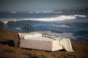 Flobeds eco-friendly mattresses sits atop bluff overlooking ocean with cresting waves; headboard is unique with wrought-iron star pattern and wood "wings" at the lower sides; bluff grasses are not tall and palisades are visible across the bay - photo
