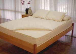 All natural non-toxic mattress with pillows on light-wood bedframe, cover is slightly folded back; dark floor; windowed wall at rear is covered in sheer draperies; side table in background holds orchids and Buddha statue - photo