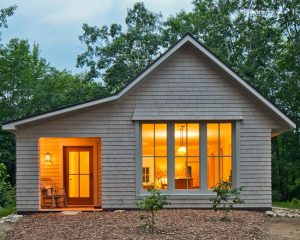 exterior view of quaint prefab house clad in clapboard in woodsy setting; warm light glows from within - photo