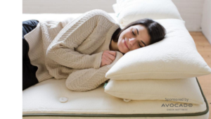 Smiling woman with dark hair dressed in beige sweater lies on mattress and has two pillows under her head, eyes closed; bright room with white walls and light wood floors. The words "Sponsored by Avocado Green Mattress" appear at lower-right - photo