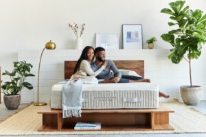 Young African-American couple cuddling on Hybrid Latex Mattress from Brentwood Home; wood bedframe, white fishes and platns decorate light-filled room - photo