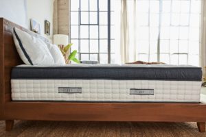 Unmade Oceano Luxury Hybrid mattress from Brentwood Home shown on wood bedstead in light-filled room; multipaned window, jute rug and plant visible - photo