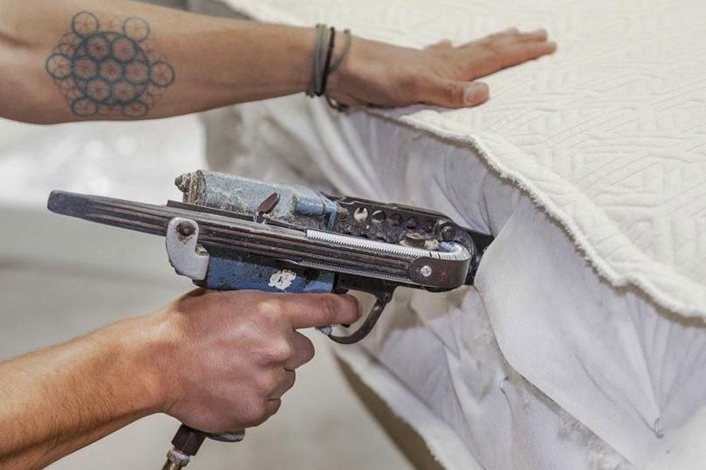 Close up of man's arms operating a pneumatic tufting gun on the side of an unfinsihed mattress