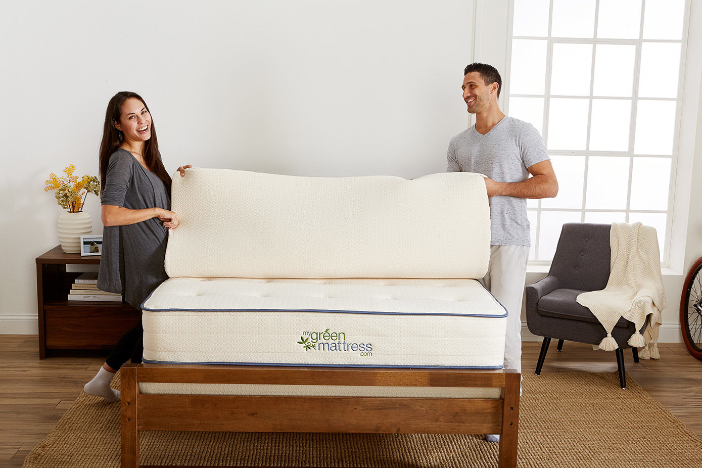 Couple layes mattess topper atop white mattress carrying My Green Mattress branding. wood bedframe stands atop textured brown rug and dark wood flooring. nightstand and gray unholstered chair in background; large white window and white wallsatop 