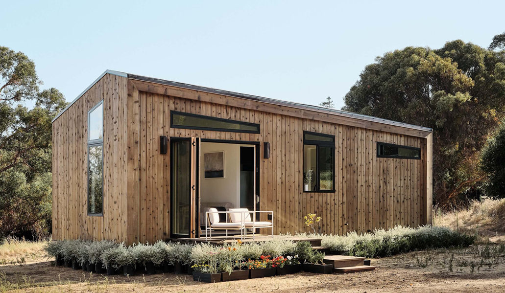 Wood-clad Abodu tiny house with open doors and seating on the desk; grass and woodland setting
