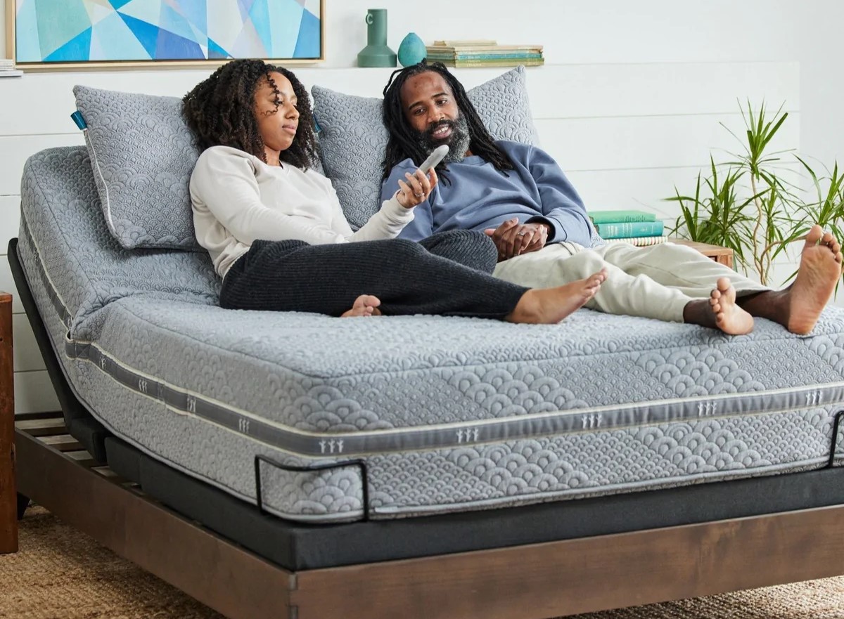 African American man with long locks and African AMerican woman with long curlswoman lie on gray adjustable mattress on adjustable base with dark wood frame; they are looking at remote control held in woman's hand.