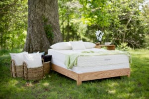 Savvy Rest Serenity Organic Mattress on light wood bedframe outside near a large tree; baskets hold fluffy pillows; light green throw complements woodland scene - photo