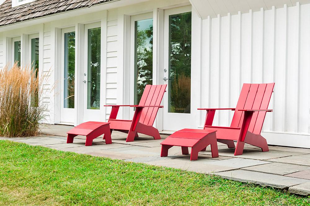 bright red Aidrondack chairs with matching footrests are set up atop flagstone patio; green lawn in foreground, white house with multiple windowed doors in background