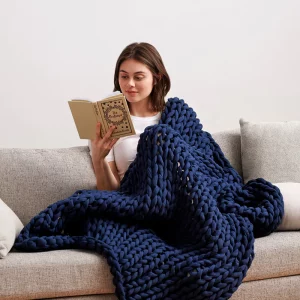 Woman sits on couch reading, covered by navy blue weighted blanket