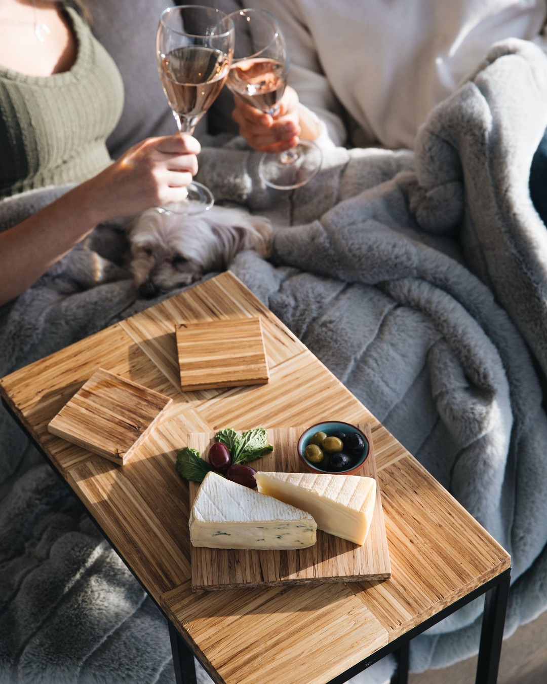 striking wood side table shows wood coasters and cheese board; two people toast with wind and dog sleeps snuggles in gray blanket