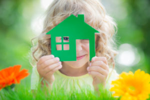 Blonde girl peers through simple cutout of house she holds in her hands; she is lying down with flowers and greenery surrounding - photo