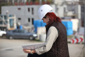 Woman in white hardhat and dark vest takes inspection notes on clipboard at construction site