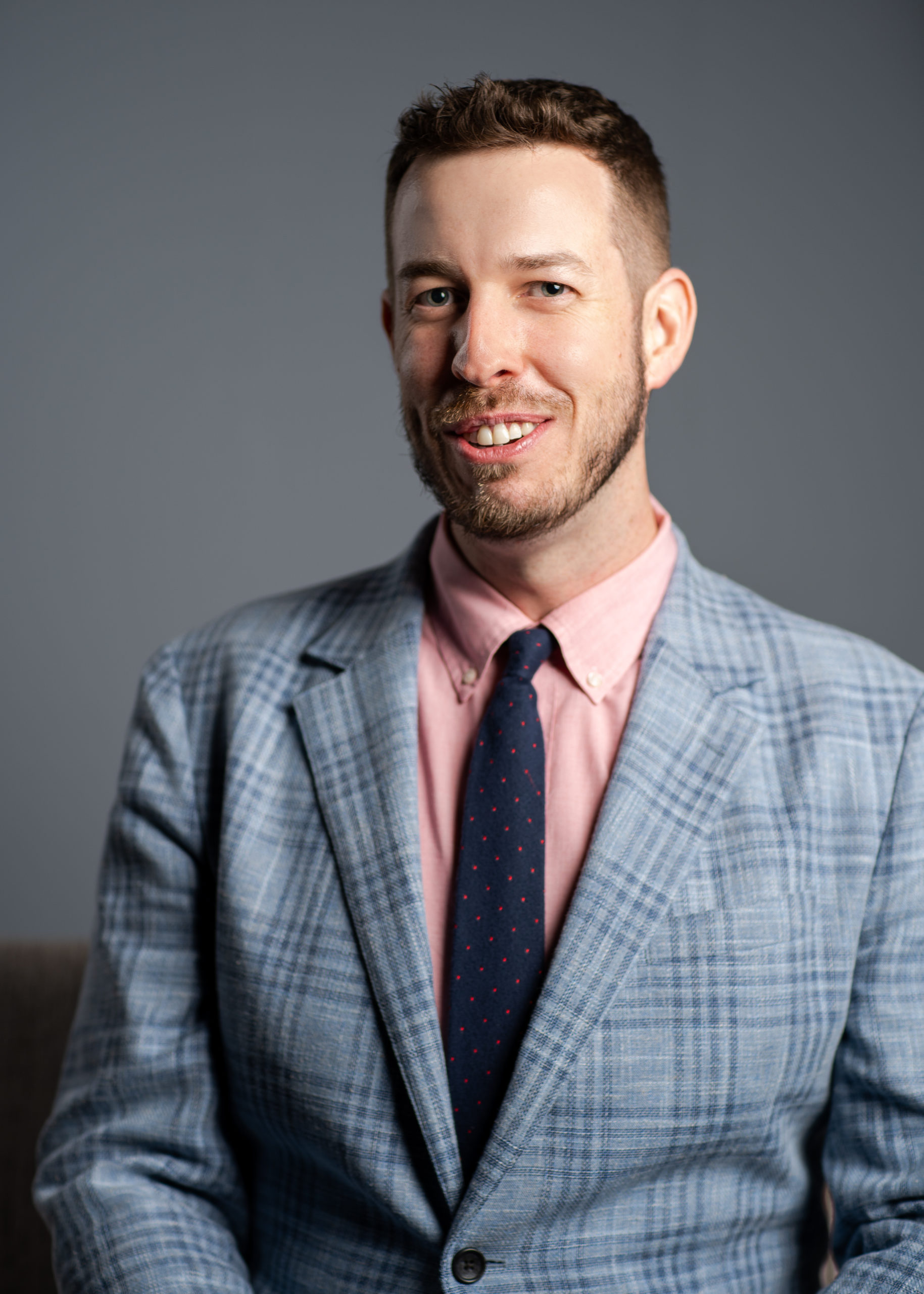 Headshot of Christopher Matos-Rogers - bearded man in gray plaid suit, drak tie and pink shirt.