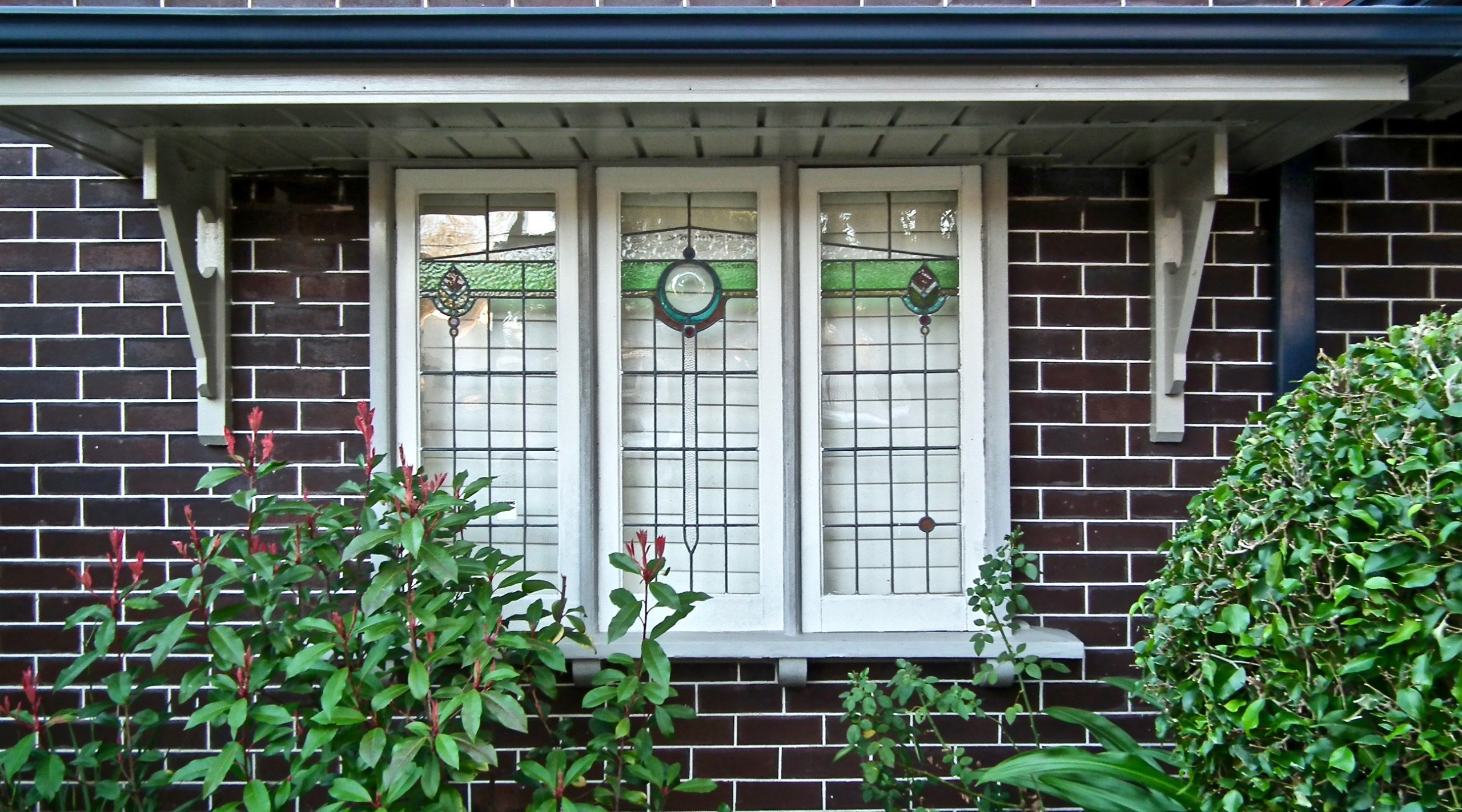 Outside of home shows dark brick with three-pane window and awning. Window has green stained glass decoration in a horizontal strip across all three windows, towards the top. Green shrubs in front of the house, covering part of the lower windows.