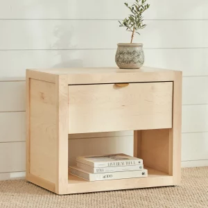 Light-colored maple night stand with small metal pull; books stacked within. Plant in gray pot on top; white eall with horizontal panels behind; beige textured rug below.