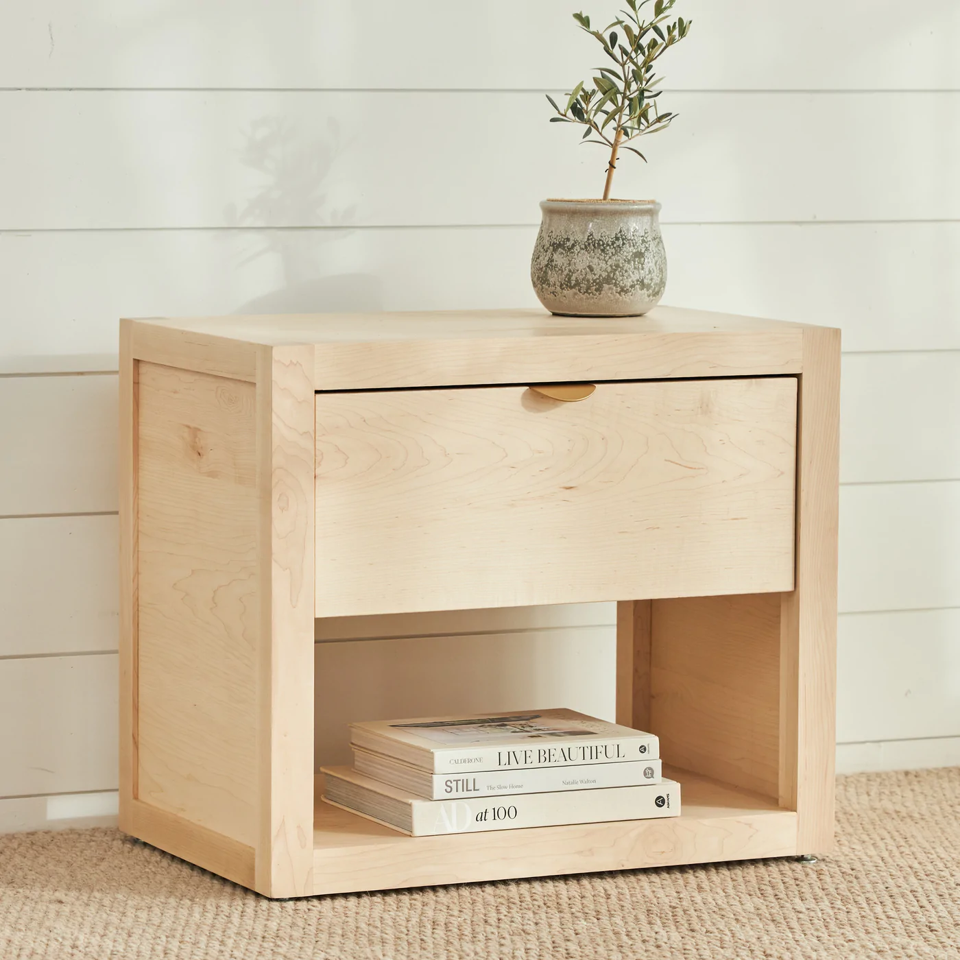 Light-colred maple night stand with small metal pull; books stacked within. Plant in gray pot on top; white eall with horizontal panels behind; beige textured rug below.