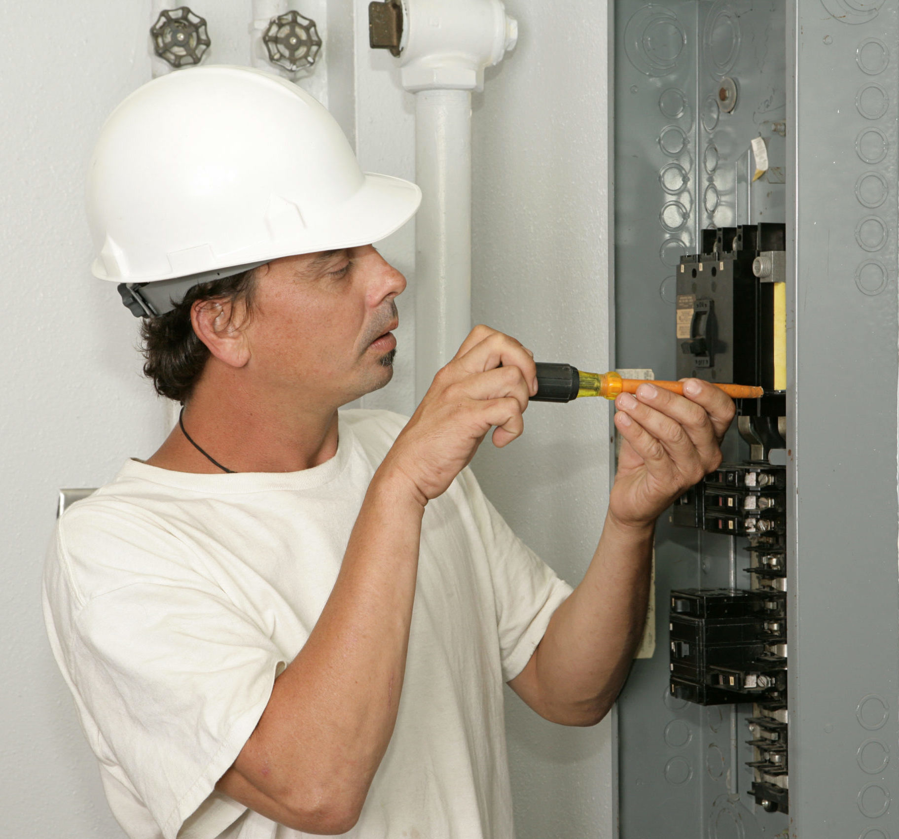Electrician in white tee and white hardhat makes adjustments on fray electrical panel
