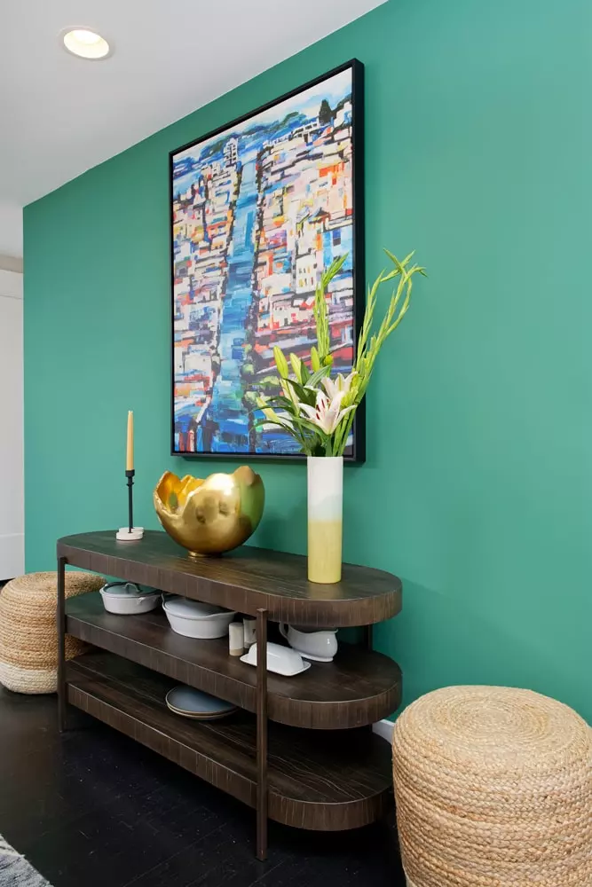 Green accent wall features colorful art, wood hall table, and straw seating/storage hassocks
