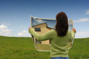 Women in green sweater facing away, green field and blue sky; she holds an large-format image of a house