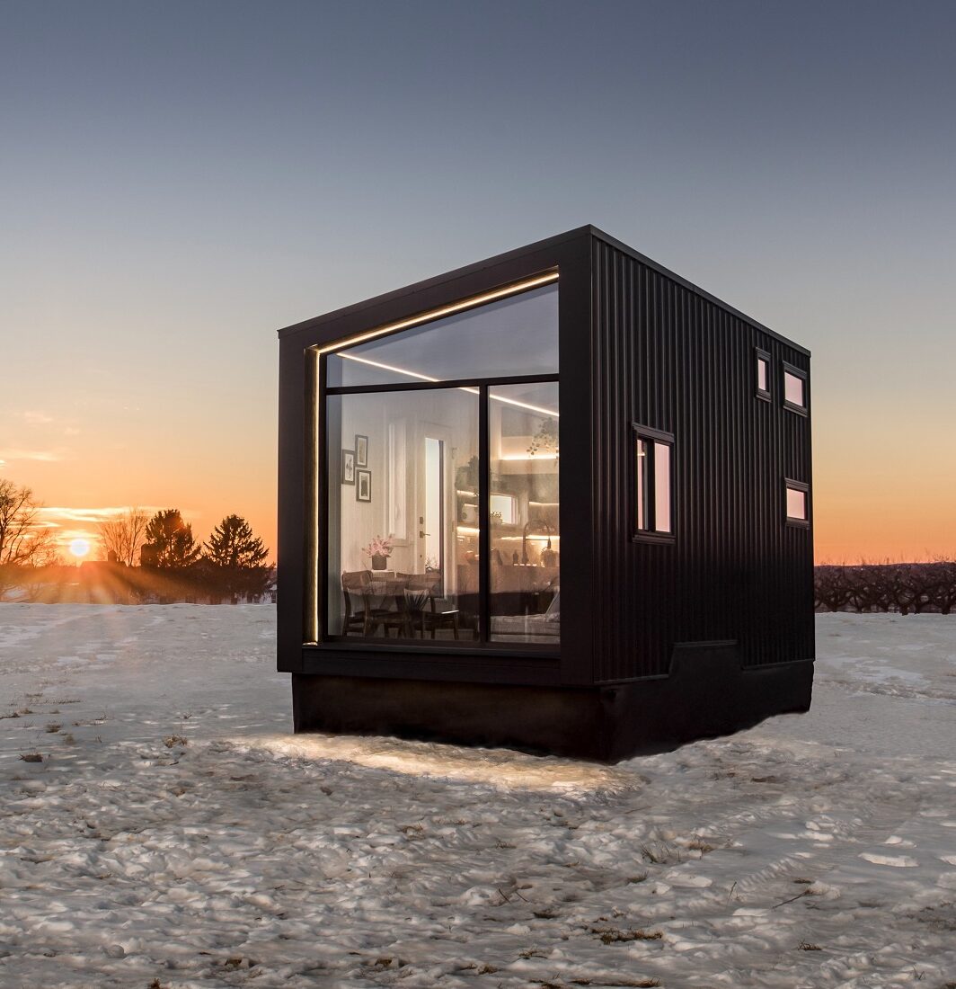 Black and glass, cube-shaped tiny home set in field of snow with setting sun,