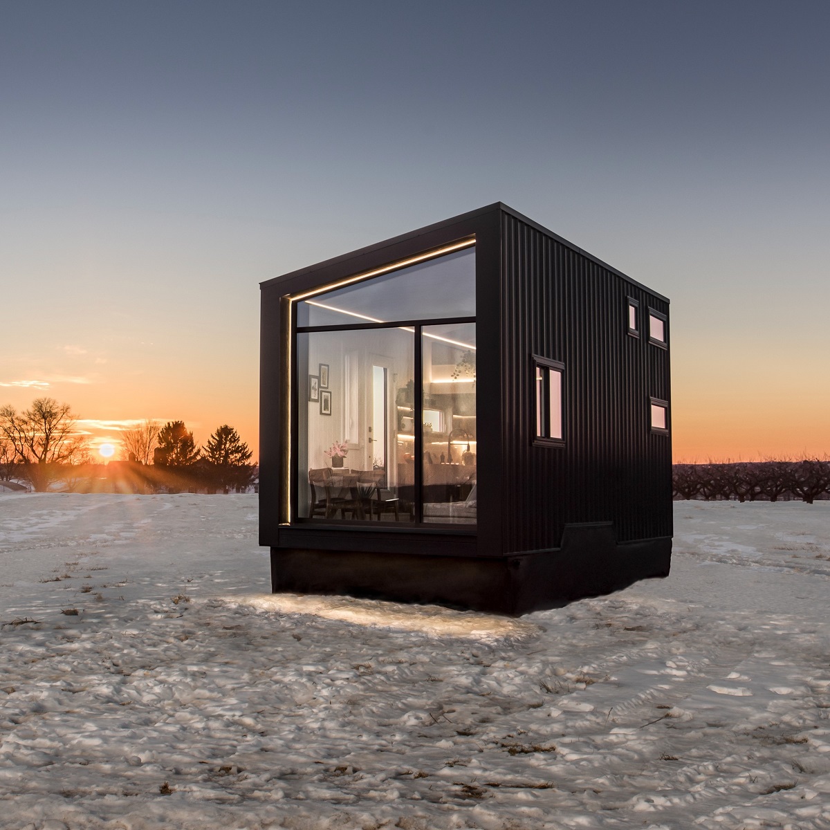 Black and glass, cube-shaped tiny home set in field of snow with setting sun,