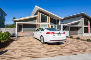 Driveway with permeable brick pavers in different shades of brown; white car with EV charging from modern house