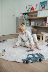 Yourng boy with blonde hair play on area rug with car images; rug lies on wood floor in bedroom/playroom with white and glray walls and white and wood furniture
