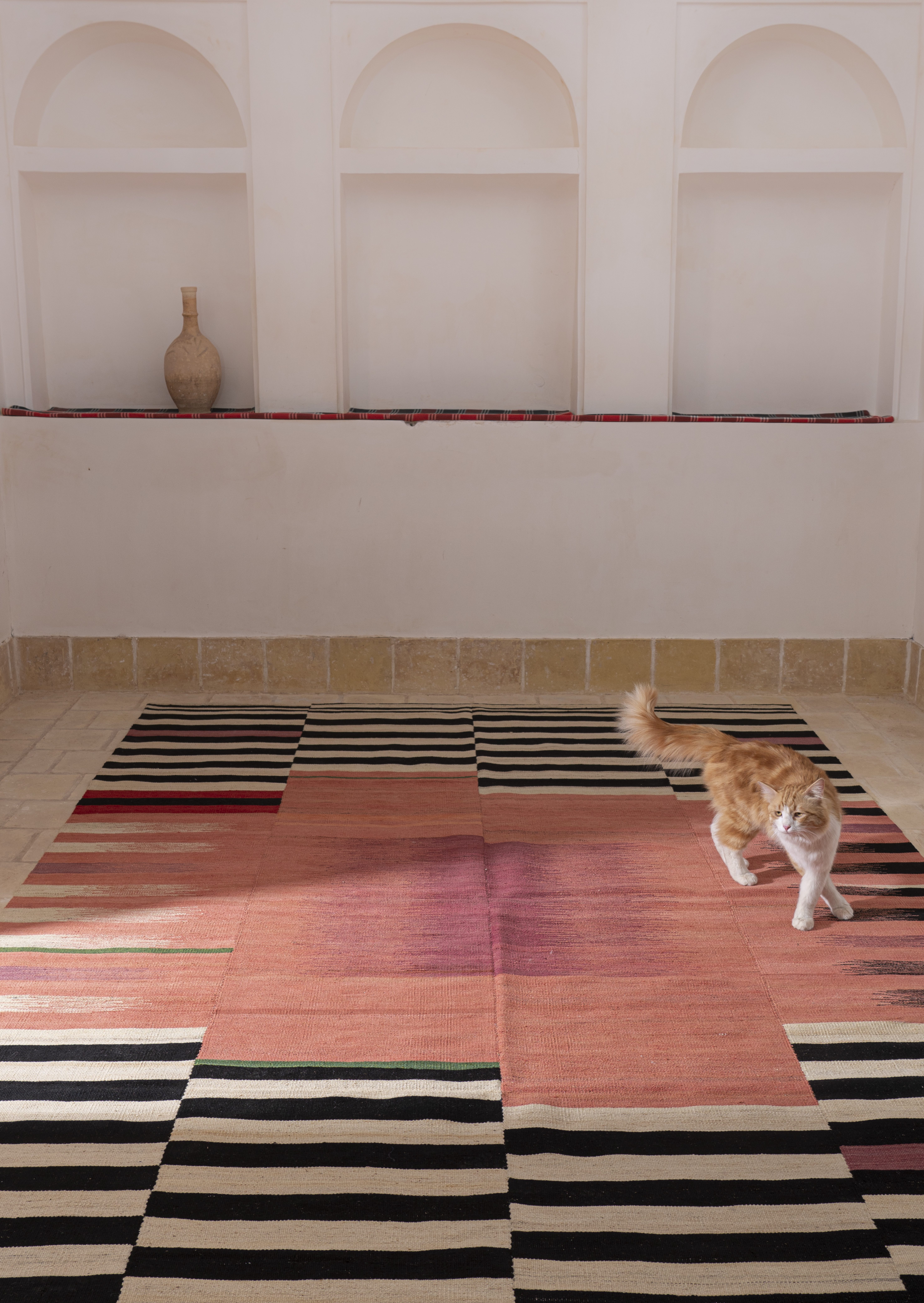 Room with white wall and tiled floor features striking area rug of beige, black, re , orange and mauve. Cat walks on rug.