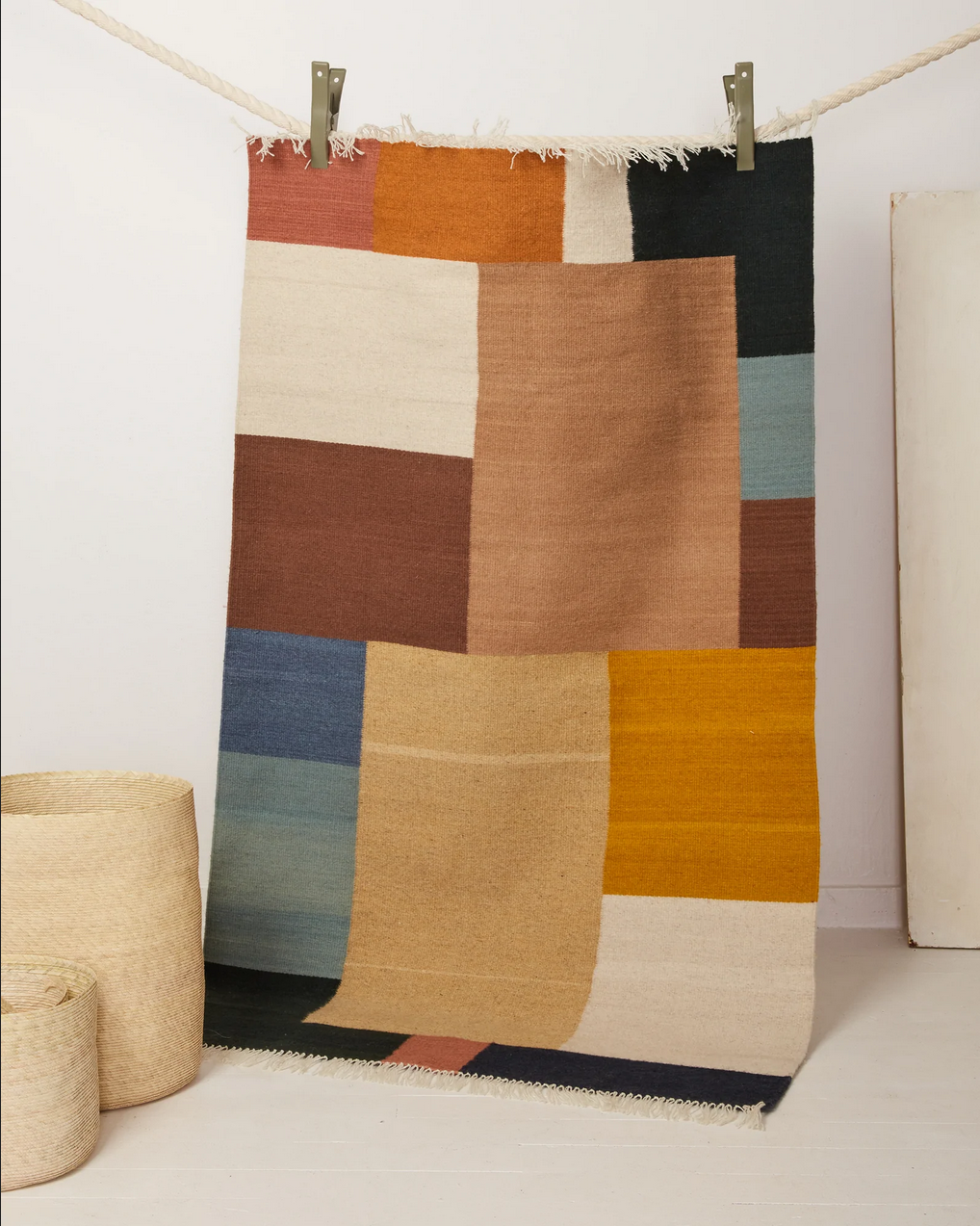 Patterned area rug in orange, brown, blue, beige with fringe; suspeced from clothesline in a white room
