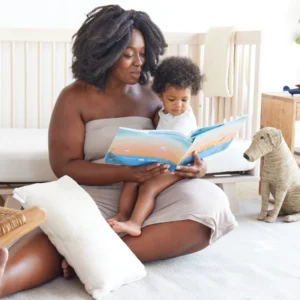Arican American mother seated on the floor in a white nursery; her infant son is on her lap and they are reading a book; stuffed dog and nontoxic toddler pillow nearby