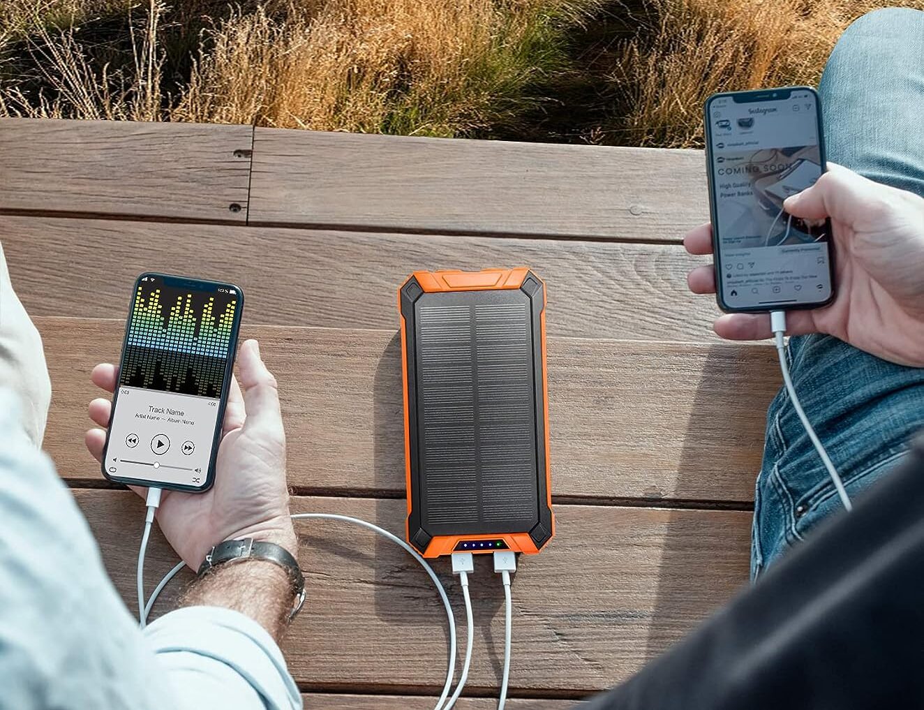 Lokking down onto bench between two seated people, organe solar battery pack charges two working phones held in the persons' hands - photo