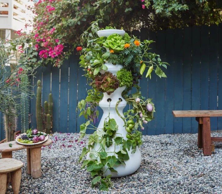 White tower with vegetables and flowers growing; approx 6 ft high; shown in graveled area of a back yard with cactus and blue wood fence - photo