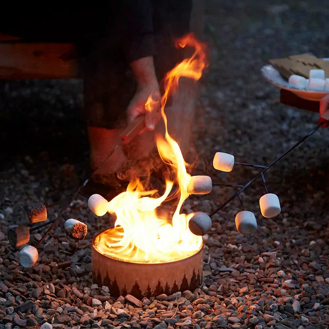 A small fire burns on a gravel surface; people's hands can be seen and numerous marshmallows being roasted on sticks - photo