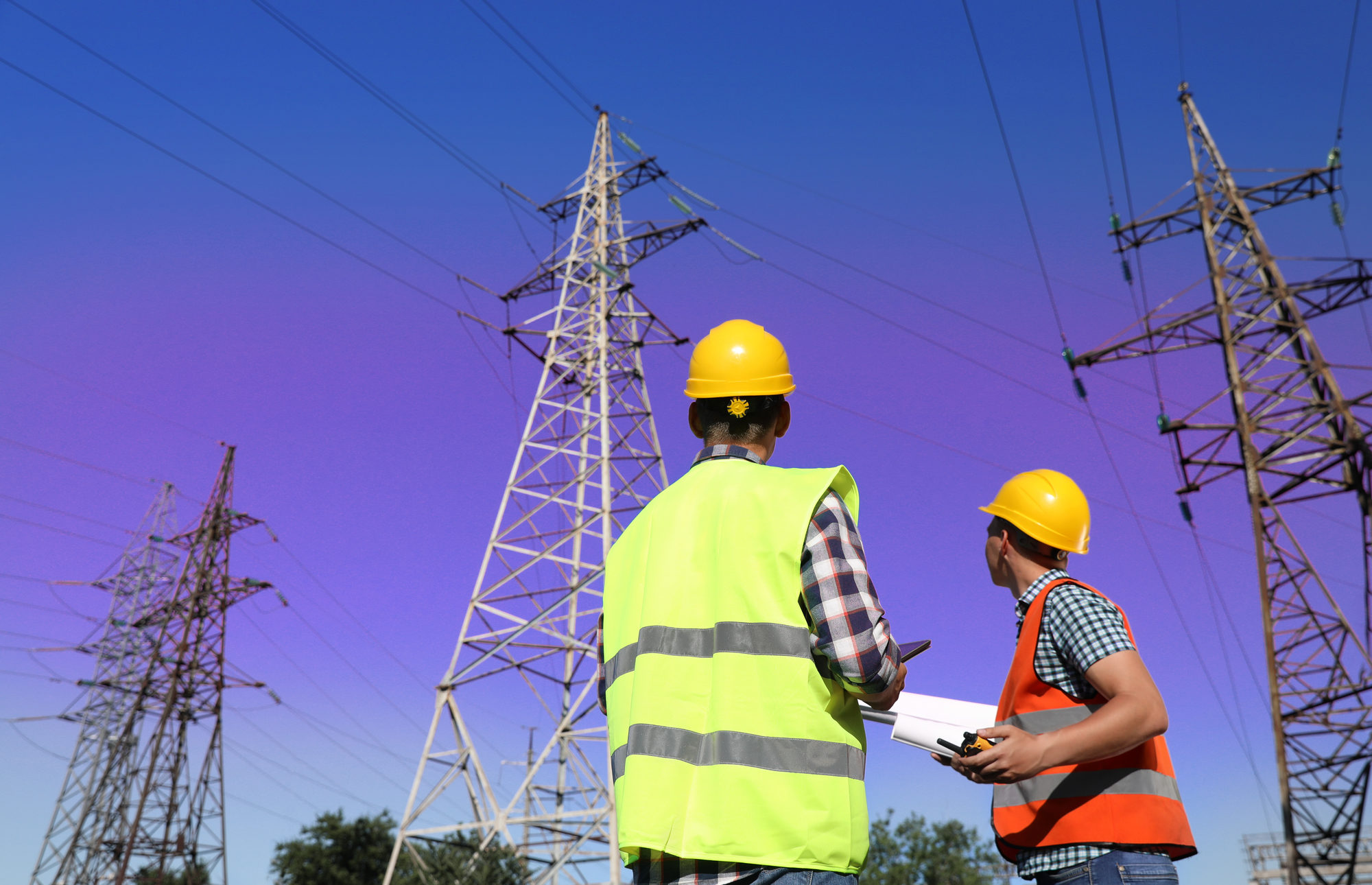 Two professional electricians in neon vests and hardhats look up at electrical towers and lines against a blue sky.