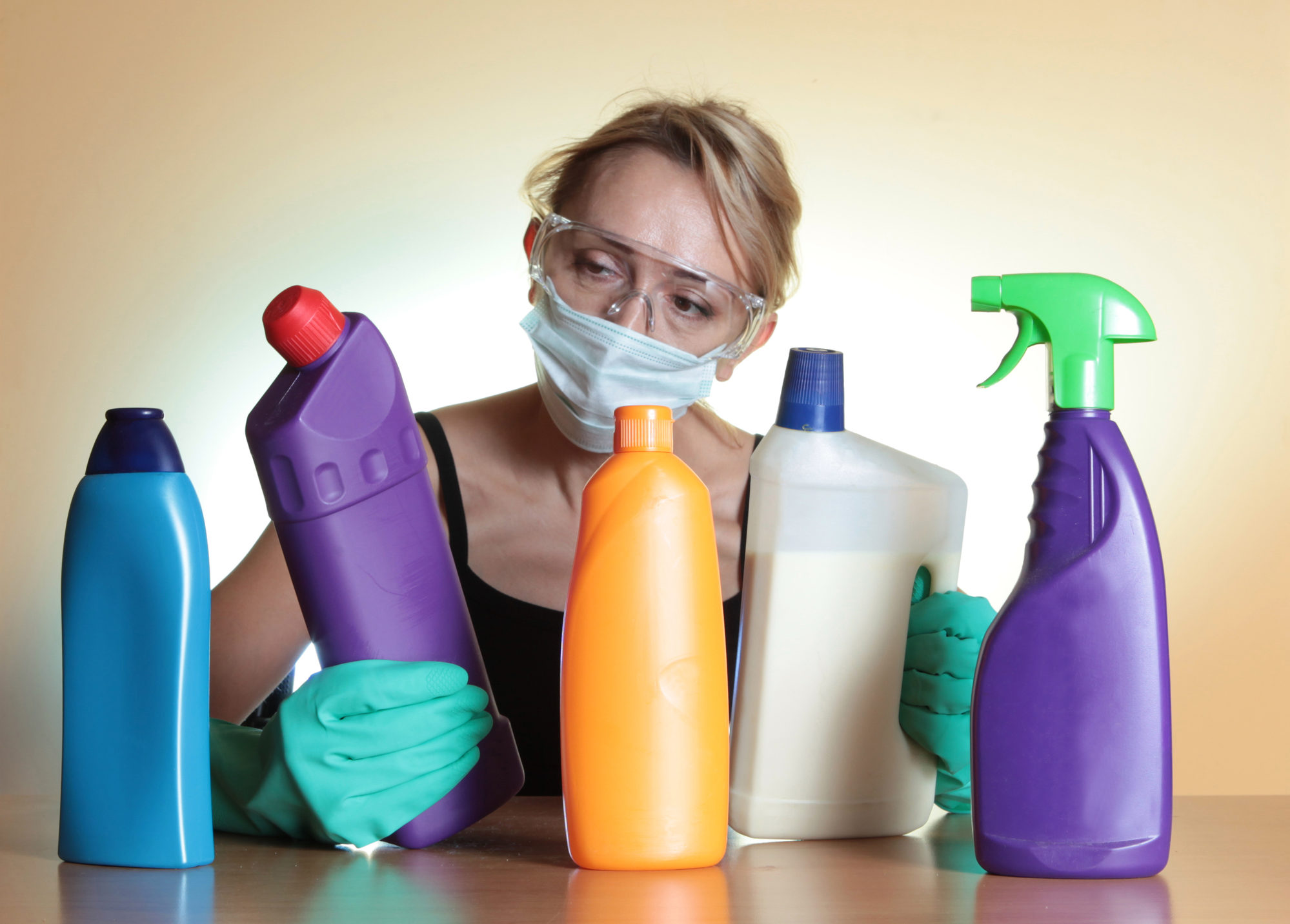 Woman in mask and goggles examines colorful bottles of toxic cleaning products,