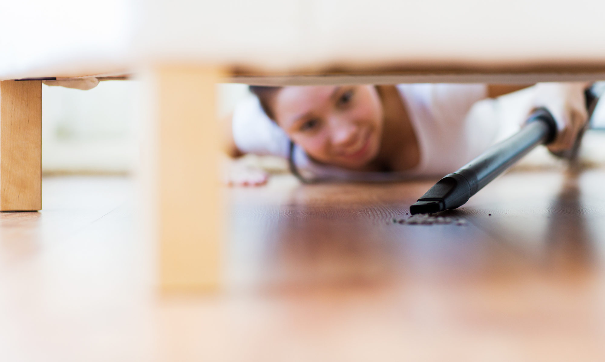 Blurred woman in background lies on floor and extends vacuum wand under furniture to pick up dust and debris