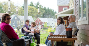 Group of adults and one child on a porch sit smiling at each other; playground and other colorful houses in the background