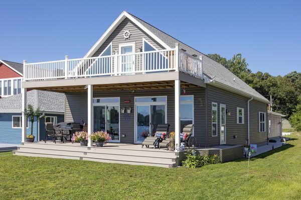 Double-height modular home with unique windows, outdoor living and dining areas on lower deck; upper deck with white vertical railing, nautical detail at apex; green lawn in foreground and waterway and sky reflected in windows