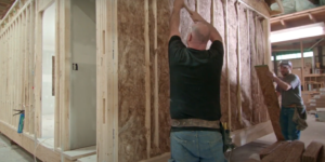 Two workers at modular home factory install insulation in wood framing.