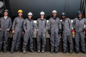 Line of industrial workers, facing the camera and smiling, wearing hardhats, work boots, and gray coveralls; standing in a line in front of large industrial container.