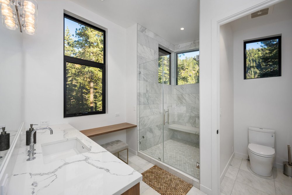 Modern bathroom with white tile and walls; white marble floors, in shower, on countertop; glass-door shower and woodlands visible through windows - photo