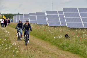 Two young people in helmets and dark jackets are biking on trail along side solar panels surrounded by grasses and wildflowers; black dog in foreground and persons walking in the rear - photo