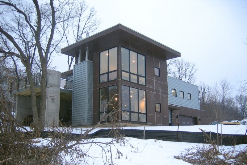 Exterior view of two-story modern home with extensive windows and multiple cladding materials; warm light shows from windows against snowy landscape and bare trees - photo