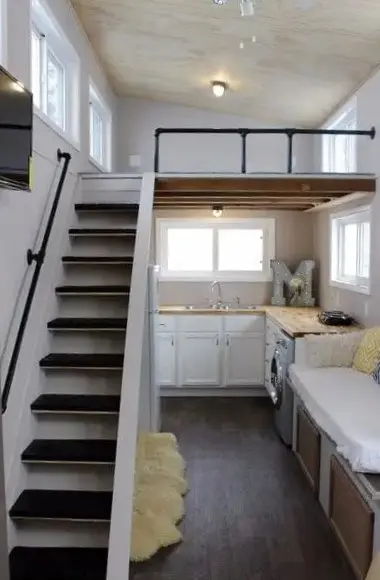 Interior view of affordable tiny home looking up stair to loft; sunny white kitchen shown