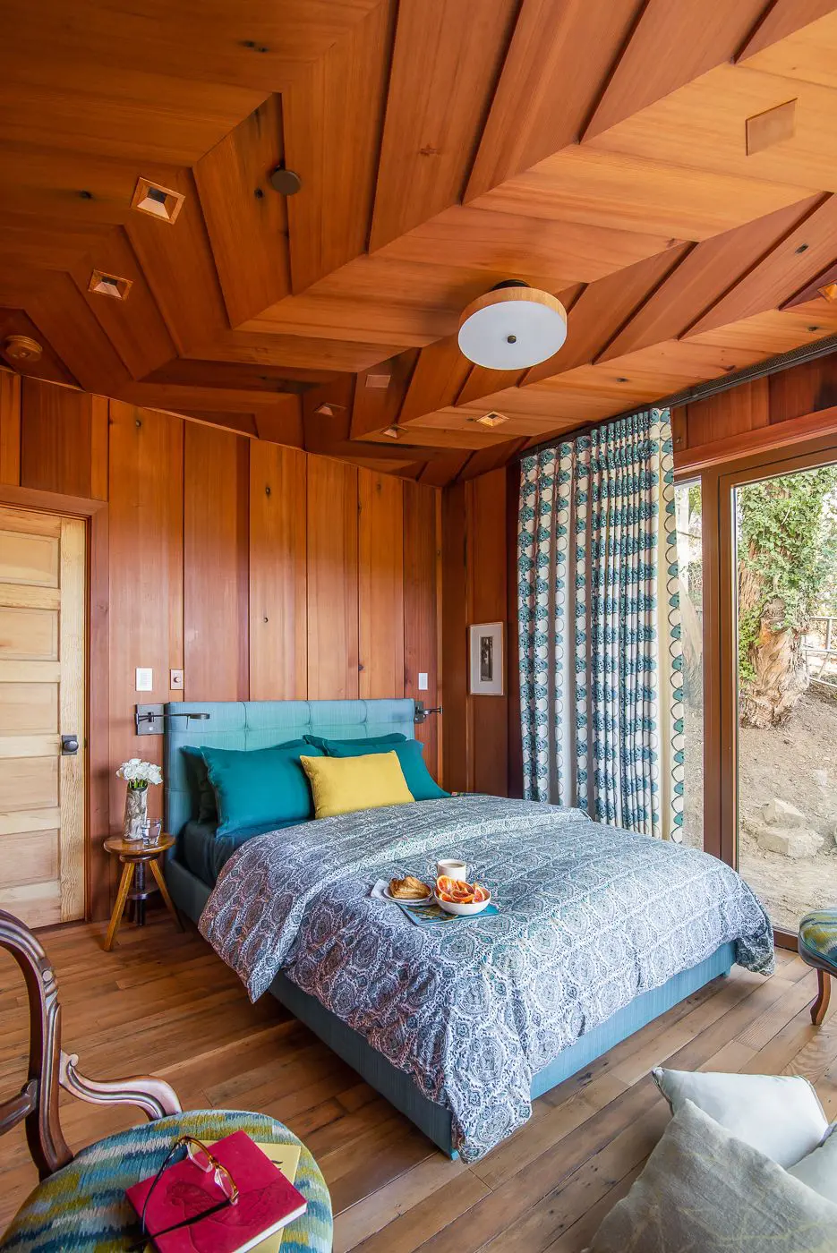 Bedroom with wood wall paneling and chevron wood ceiling and colorful bedspread; floor to ceiling curtains on sliding door to outside - photo