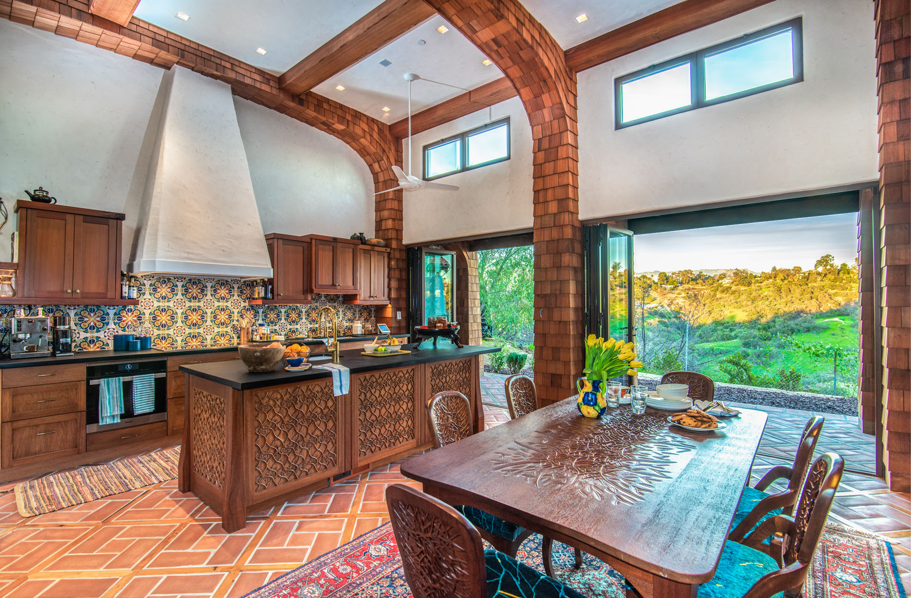 Open plan kitchen and dining area with intricate carving on island and table/chairs; large open accordion doors open onto year - photo