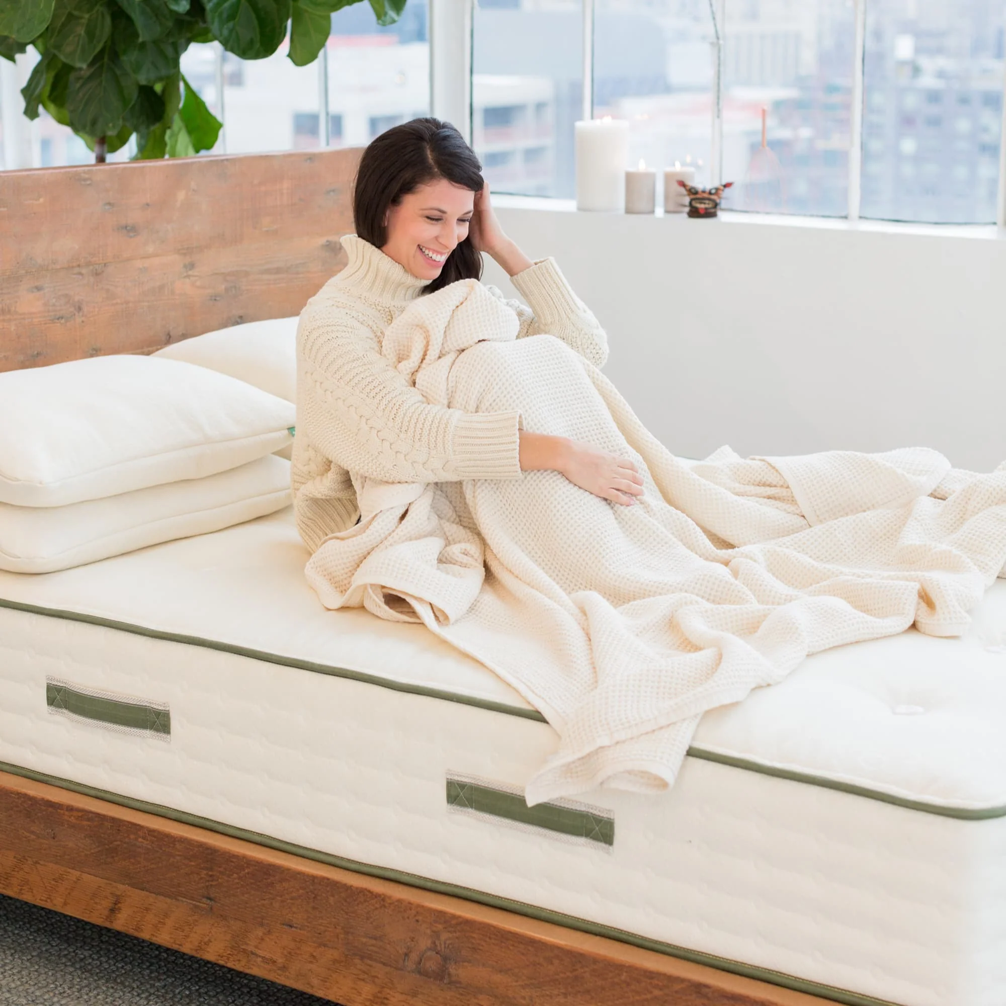 Woman sitting up on eco-freindly Avocado mattress in light-filled high-rise room; wood bedstead and plant visible - photo