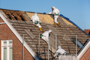 Workers in white hazmat suits and respirators are removing material from residential roof; asbestos hazard - photo