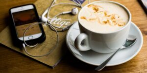closeup of tabletop holding white cup of coffee latte, and smartphone displaying podcast with wired headphones on brown-covered design-type catalog - photo