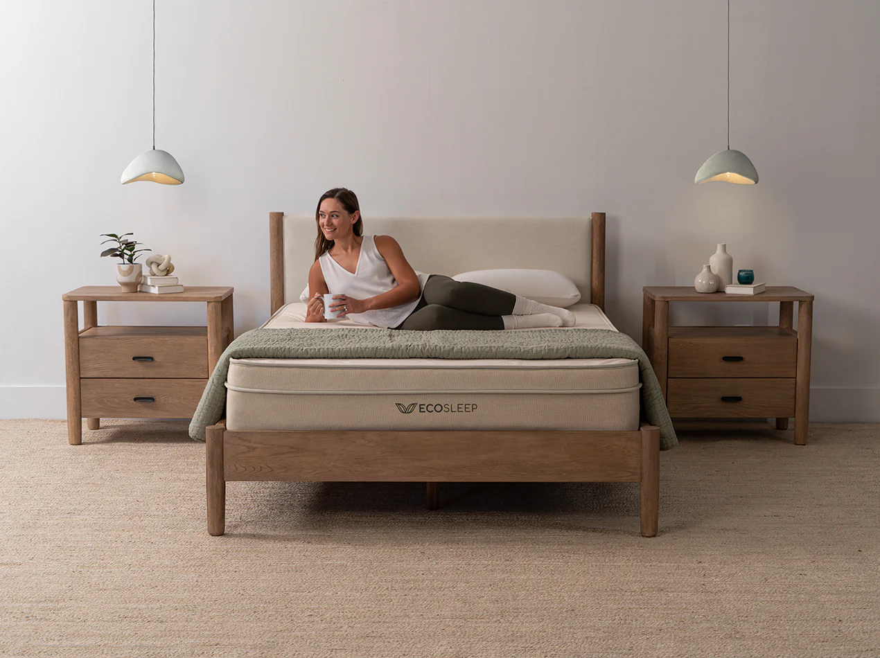 Young woman with long dark hair rests on organic mattress labeled EcoSleep; light green quilt, tan rug, and white back wall complement minimalist wood bedstead and end tables plus two interesting white pendant lights; woman smiles, gazing off to her right - photo